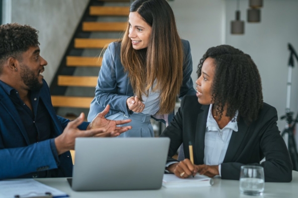 In a modern office, three colleagues - a Black man, white woman and Black woman - huddle at a laptop while collaborating on work..